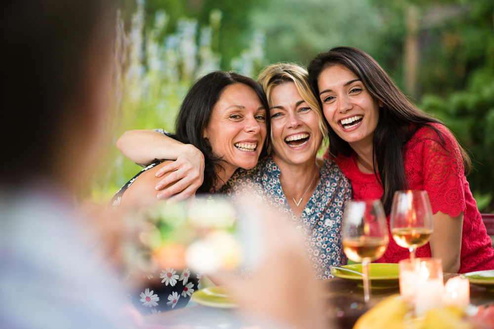 Three beautiful women posing for a photo around a terrace table by a summer evening.
